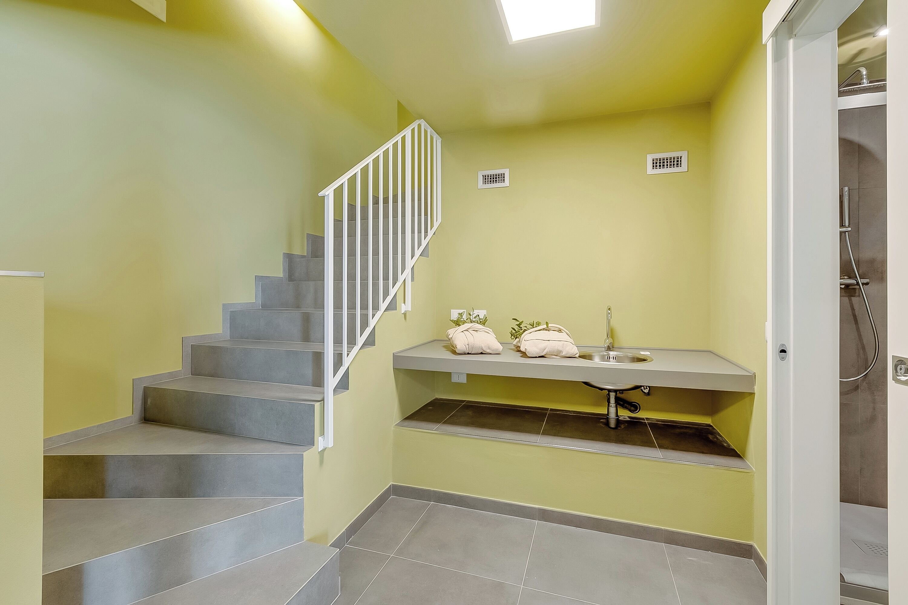 Ein Badezimmer mit grau gefliestem Boden, einer weißen Treppe mit Geländer, einem Waschbecken mit Spiegel und einer Duschkabine.