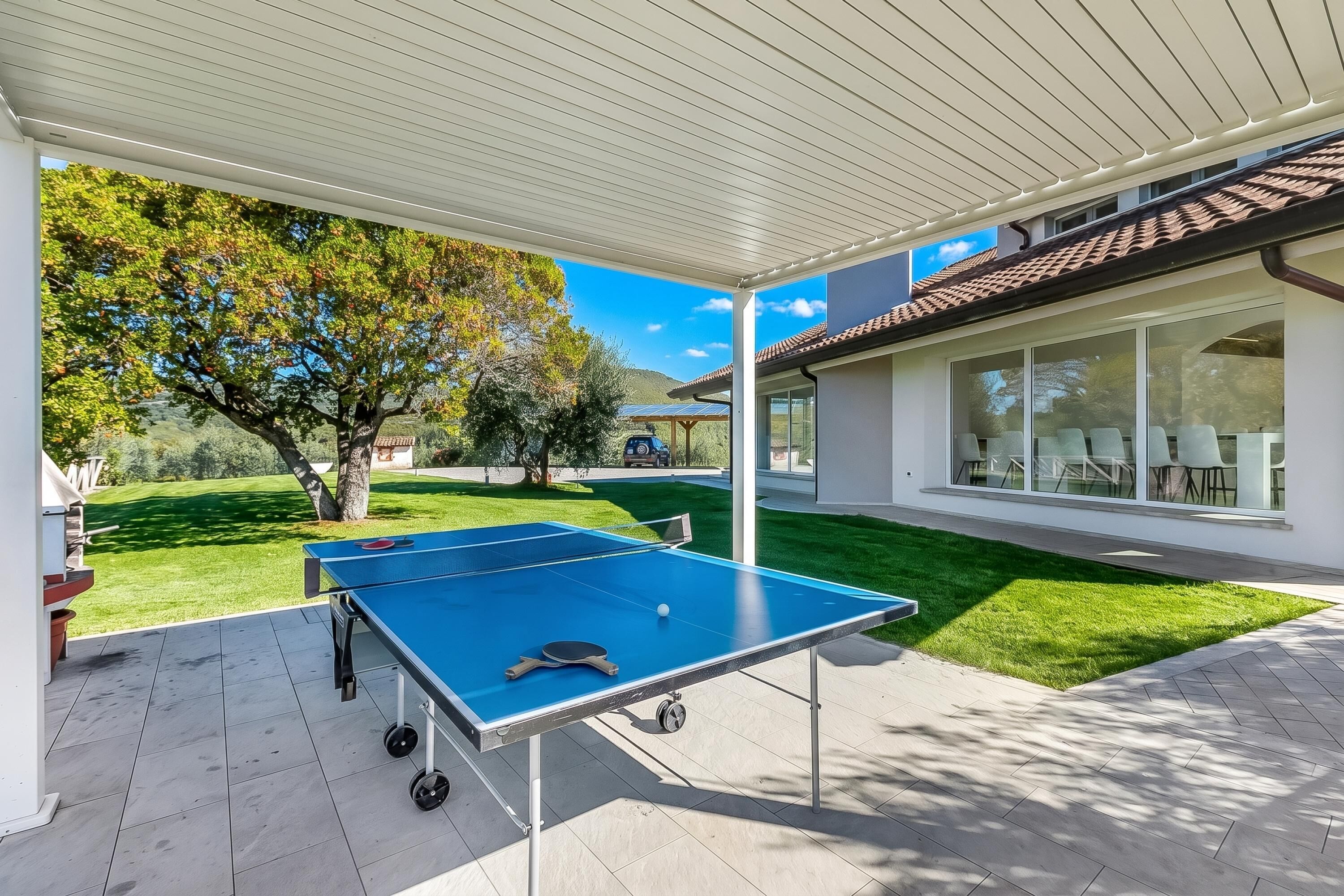 Ein Tischtennis-Tisch im Freien auf einer überdachten Terrasse mit Blick auf ein Haus und eine Rasenfläche.
