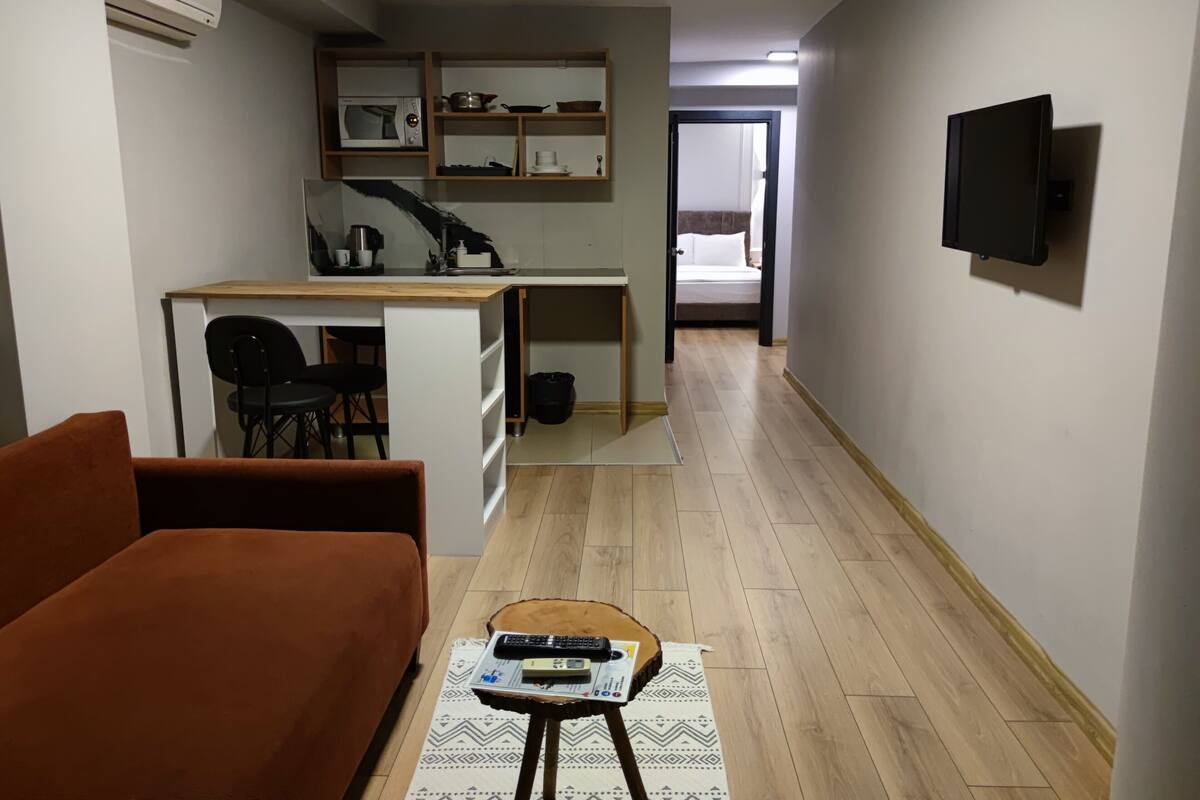 A compact living space with a kitchenette, a sofa, a coffee table with remote controls, and a television mounted on the wall.
