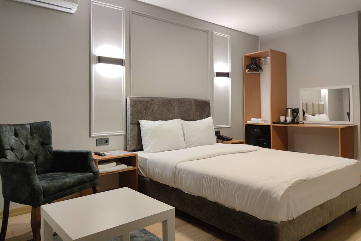A hotel room with a bed, a chair, a table, a nightstand, a wardrobe, a mirror, and an air conditioning unit.