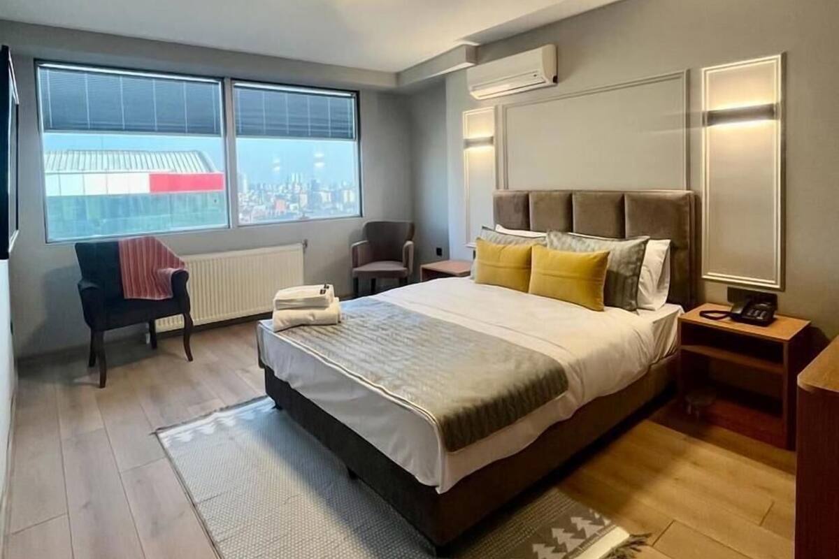 A modern hotel room with a large bed, a chair, a desk, and a window with a city view.