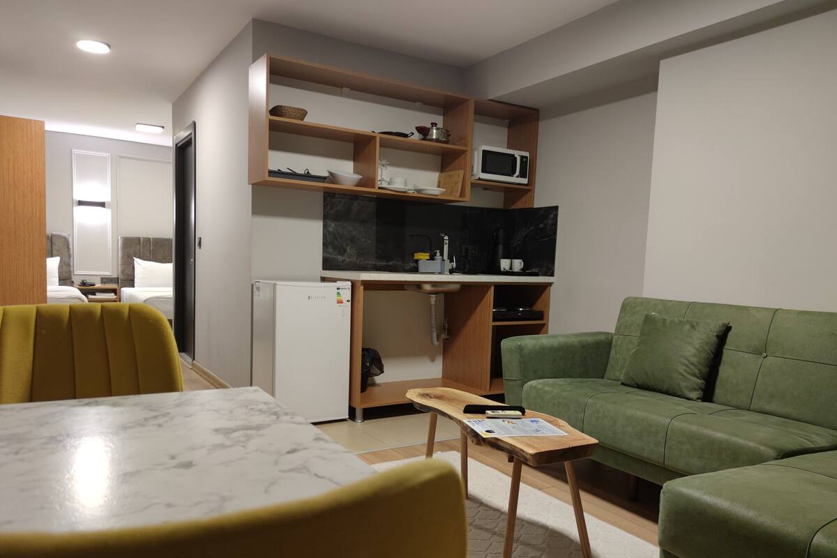 A compact living space with a dining table, a green sofa, a small wooden side table, and a kitchen area with a microwave and shelves.