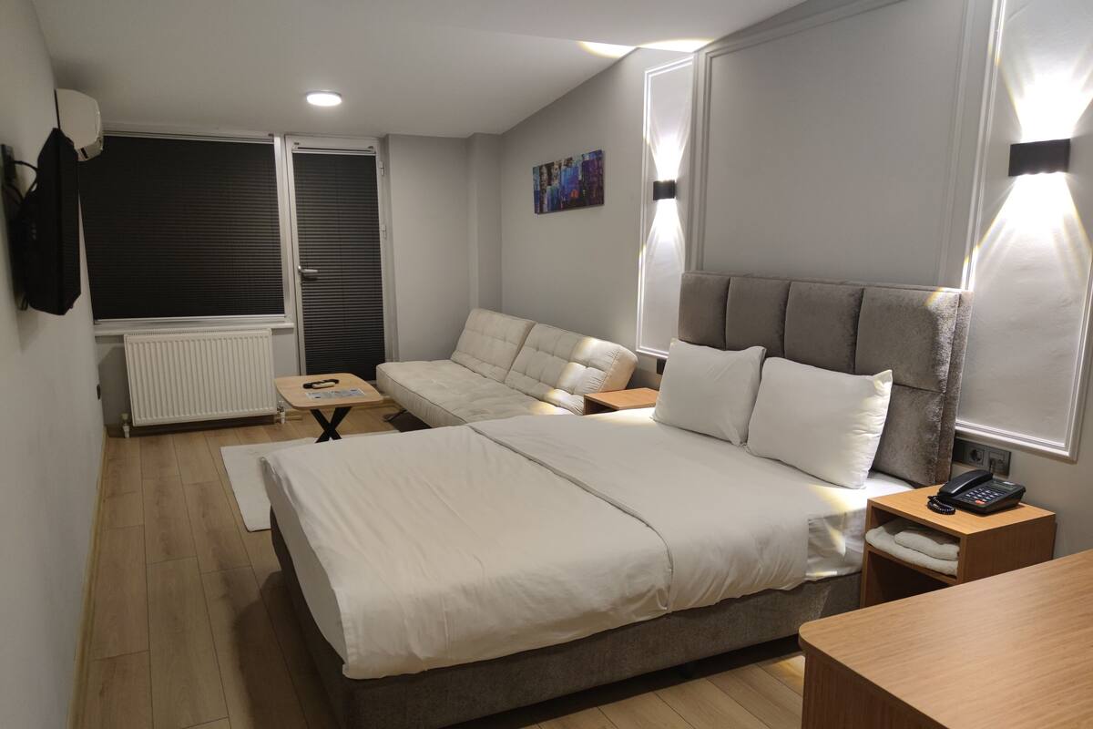 A modern bedroom with a large bed, a sofa, a small table, and a bedside table with a phone and a hairbrush.