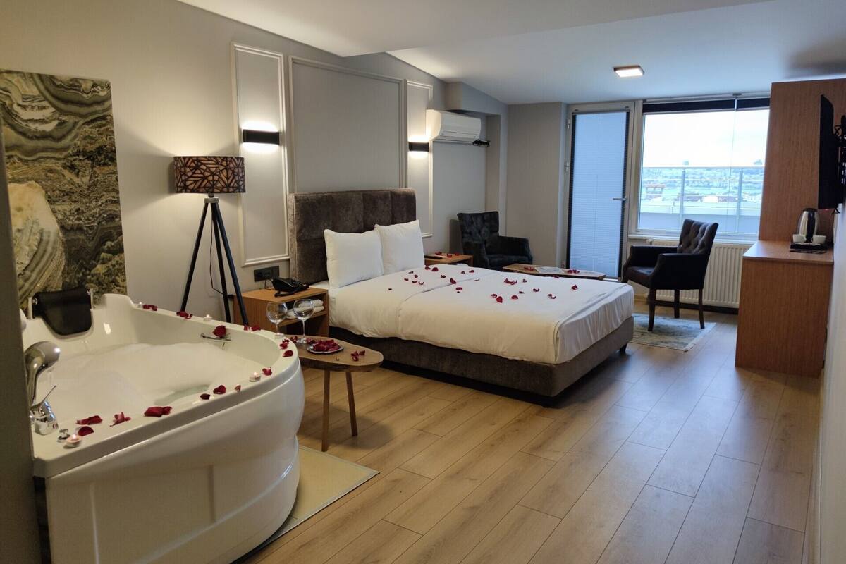 A hotel room with a large bed, a sitting area with a chair, a desk, a bathtub with rose petals, and a city view through the window.