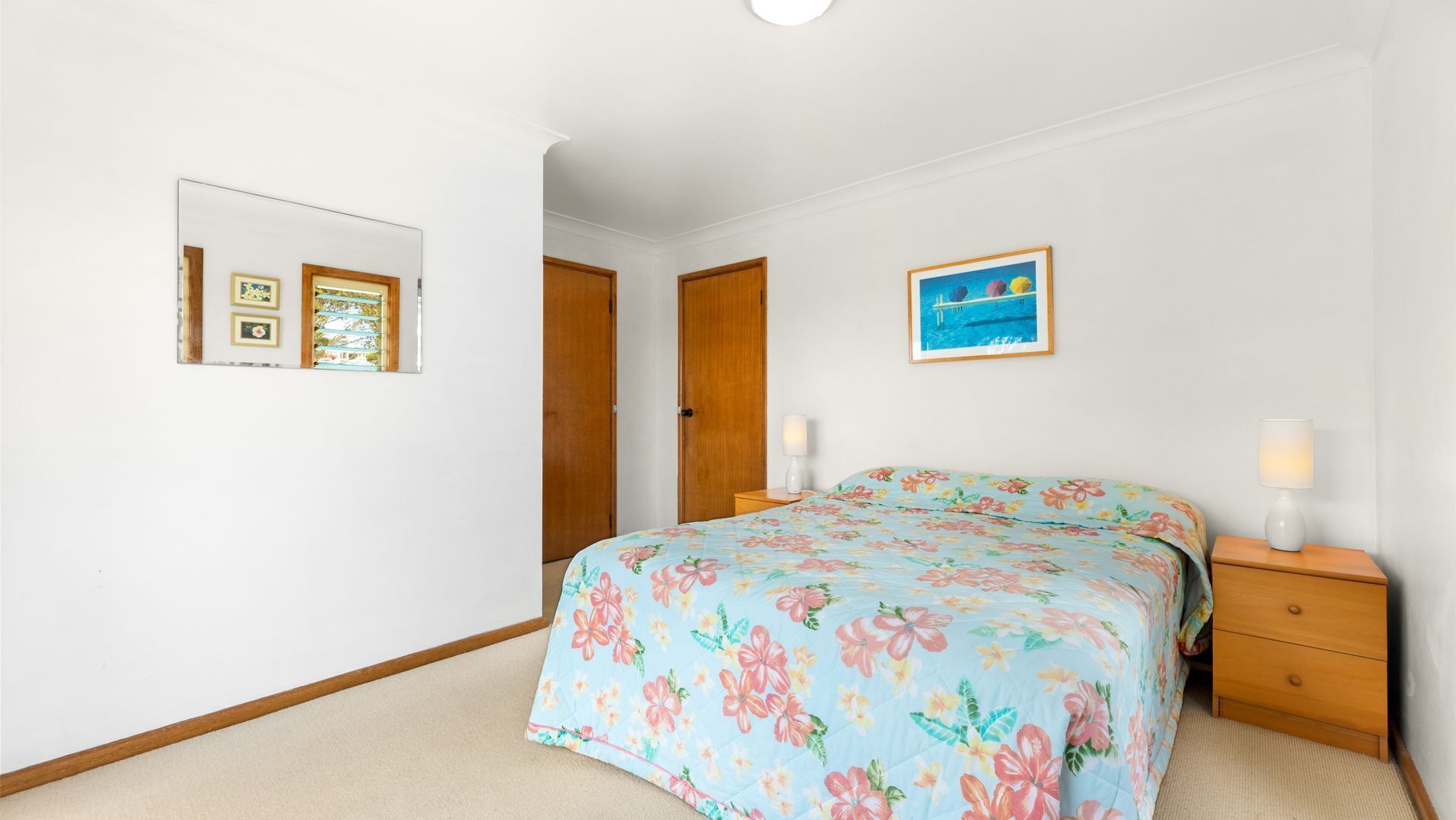 Een slaapkamer met een bloemenprint beddeken, een bedlampje, een ingelijst beeld op de muur en een raam met zonwering.