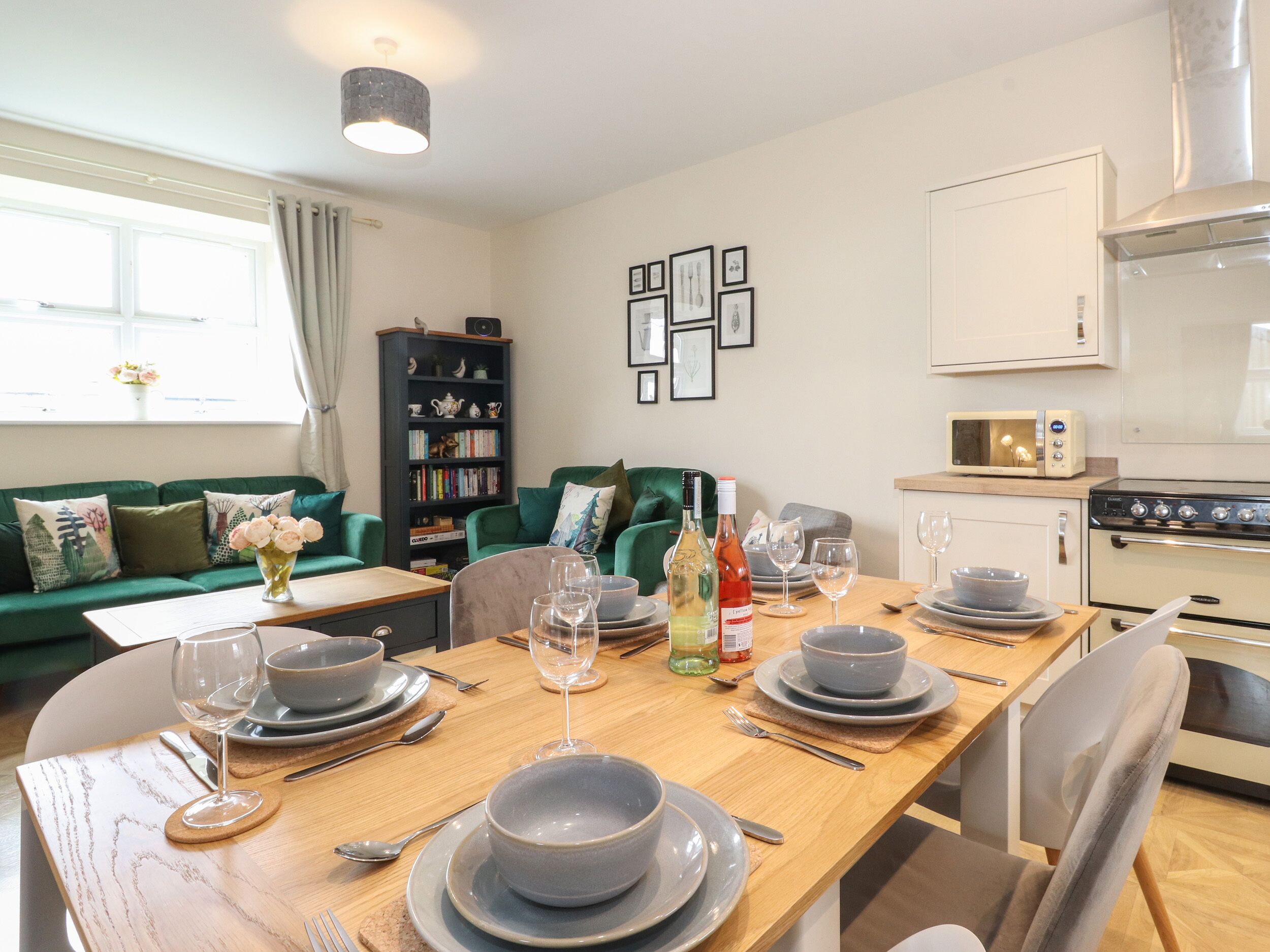 Un coin repas avec une table en bois dressée pour quatre personnes, entourée de chaises. On aperçoit un canapé vert et une bibliothèque à l’arrière-plan.