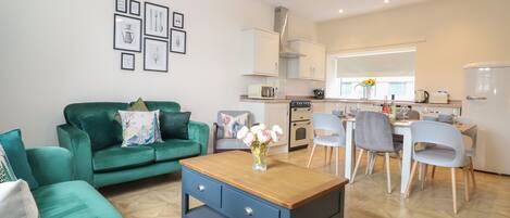 A modern living room with a green sofa, a wooden coffee table, and a dining area with a white table and grey chairs.