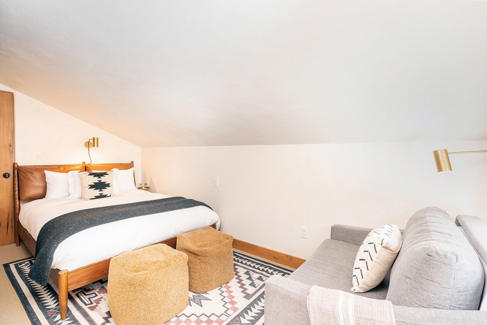 A bedroom with a bed, two ottomans, a gray sofa, and a patterned rug.