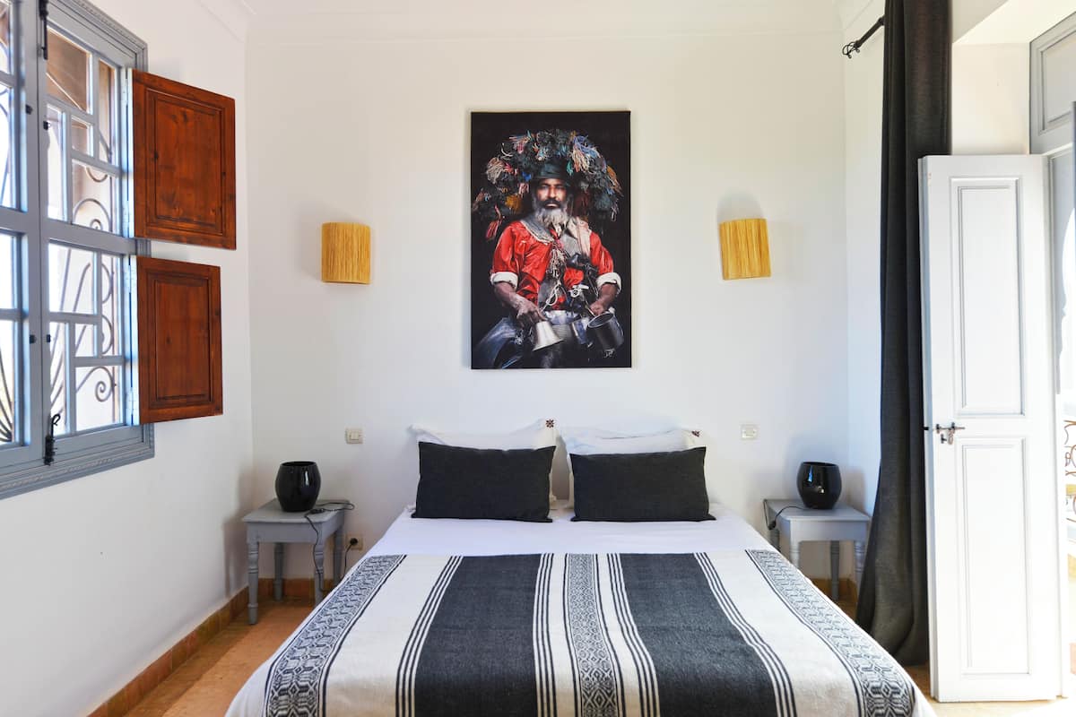 A bedroom with a bed, two bedside tables, a wall-mounted painting, and a window with wooden shutters.
