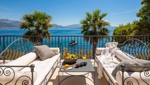 A balcony with two white cushioned sofas, a small table with a fruit bowl and a book, and a view of the sea and mountains.