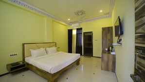 A hotel room with a large bed, a wooden nightstand, a flat-screen TV mounted on the wall, and a small desk with a mirror. - Shiv Ashray Resort (Nathdwara)