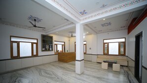 A spacious room with marble flooring, a wooden reception desk, and a seating area with a bench and ottoman. - Shiv Ashray Resort (Nathdwara)