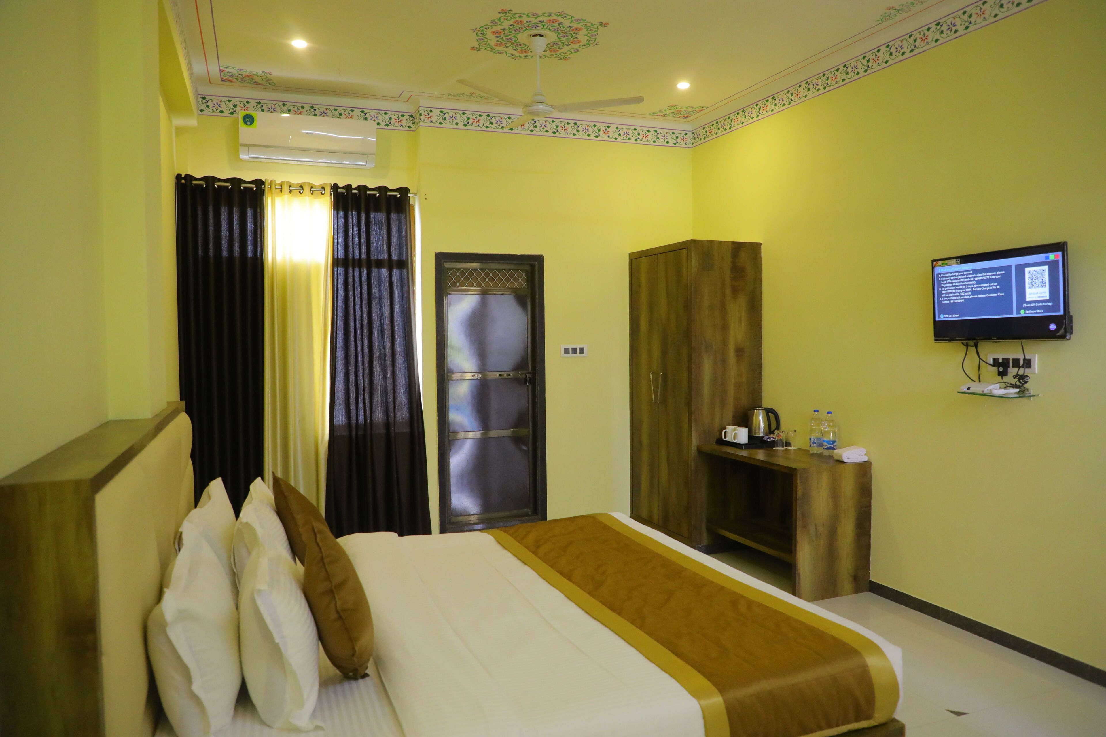 A hotel room with a bed, curtains, a television, a wardrobe, and a desk with a kettle and bottles.
