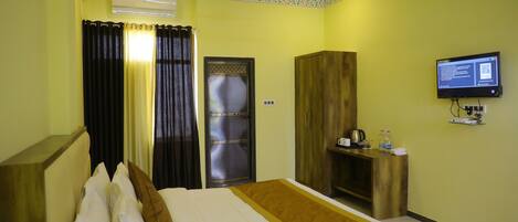 A hotel room with a bed, curtains, a television, a wardrobe, and a desk with a kettle and bottles.