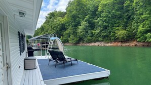 A boat deck with a covered area, two lounge chairs, a ladder, and a covered seating area with a view of a forested riverbank. - Floating Cabin - Another Day in Paradise at Whitman Hollow Marina (LaFollette)