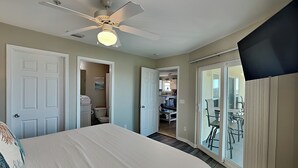 Quarto com cama, ventilador de teto, banheiro visível por uma porta aberta e porta de vidro corrediça que dá para uma varanda.