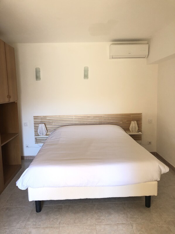 A bedroom with a bed, wooden headboard, bedside table, lamp, and a wall-mounted air conditioner.