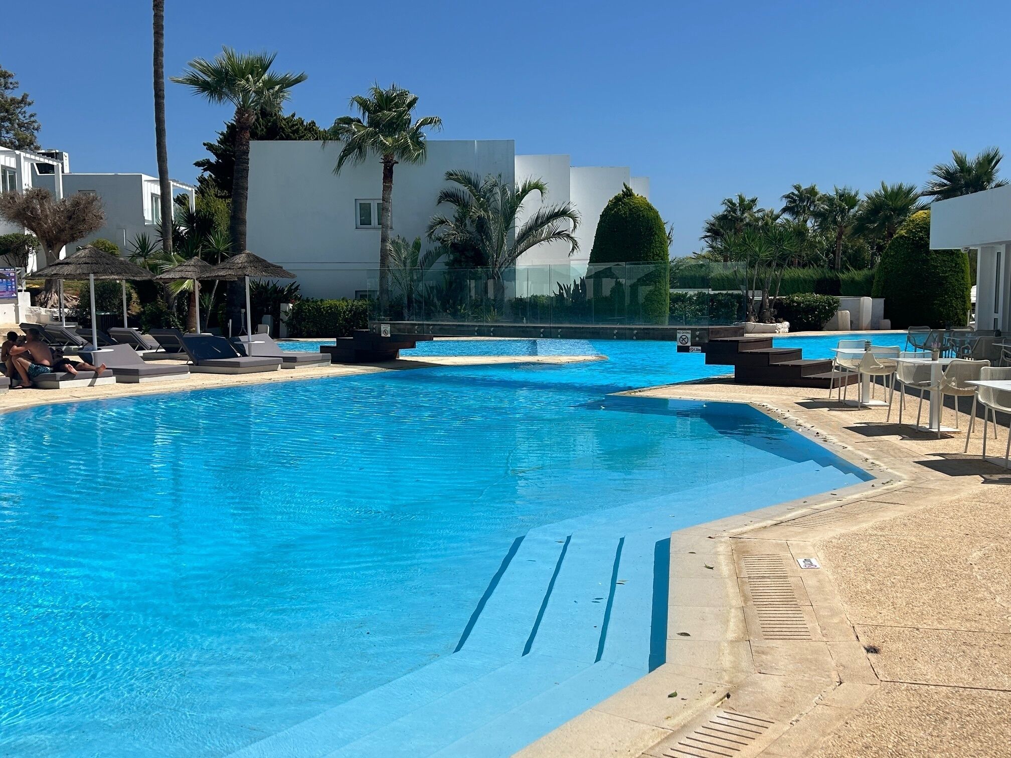 A swimming pool with lounge chairs and umbrellas, surrounded by palm trees and modern buildings.
