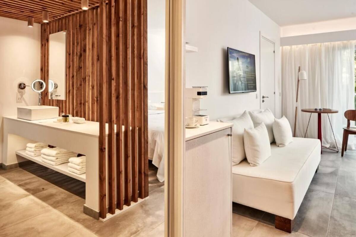 A modern hotel room with a wooden partition, a white sofa, a small table, a painting on the wall, and a view through the window.