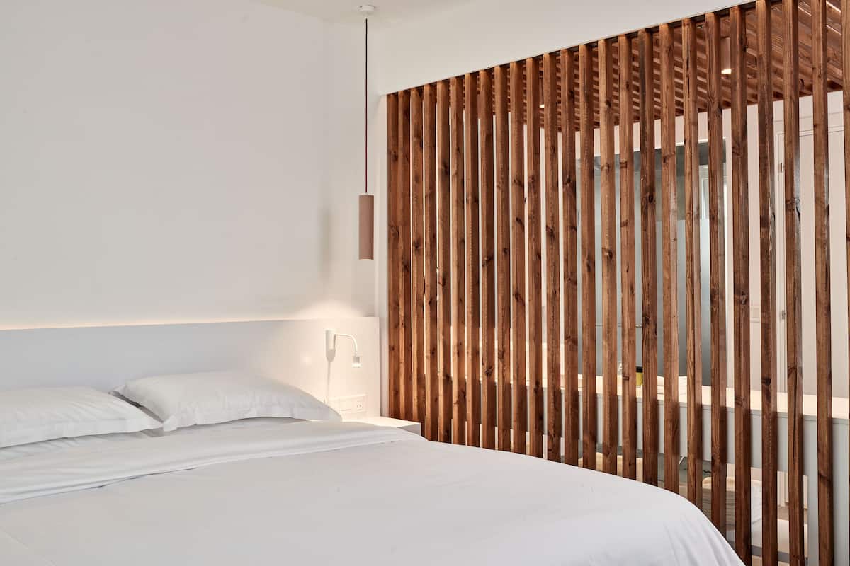A modern bedroom with a wooden slat partition, a bed with white linens, and a pendant light.