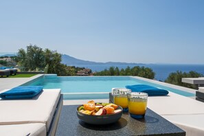 Ein Tisch am Pool mit einer Obstschale und zwei Gläsern Orangensaft, mit Blick auf das klare blaue Meer und ferne Berge.