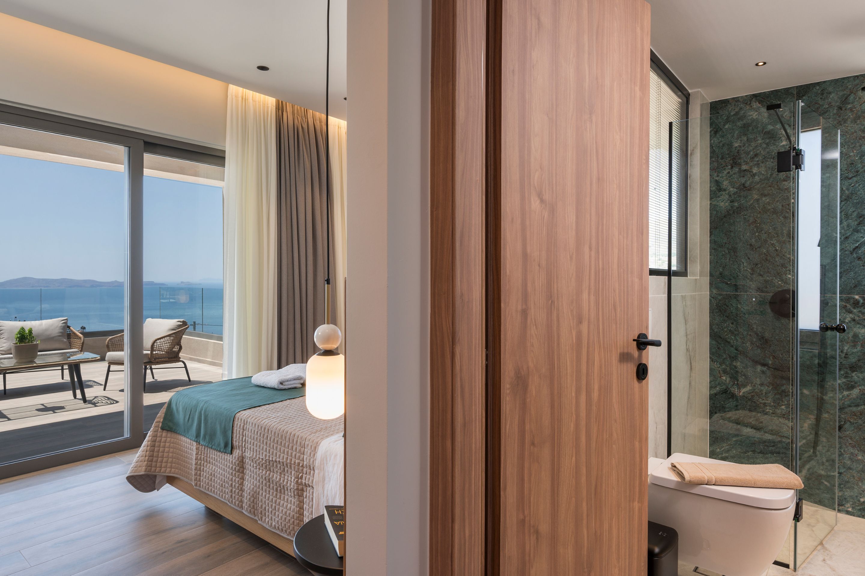 A modern hotel room with a large bed, a balcony with a view, a shower with a glass door, and a wooden door.