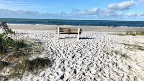 Een strand met een houten bankje en een stoel, een heldere lucht en de oceaan op de achtergrond.