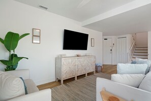 Een woonkamer met een flatscreen televisie aan de muur, een witte bank, een houten kast en een trap die naar een andere kamer leidt.