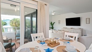 A dining area with a glass door leading to an outdoor patio, a television mounted on the wall, and a white sofa in the background. - Hidden Pearl 2 Bedroom Home by RedAwning (Holmes Beach)