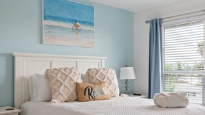 A hotel room with a bed, a painting of a beach scene, a nightstand, a lamp, and a window with curtains. - Hidden Pearl 2 Bedroom Home by RedAwning (Holmes Beach)