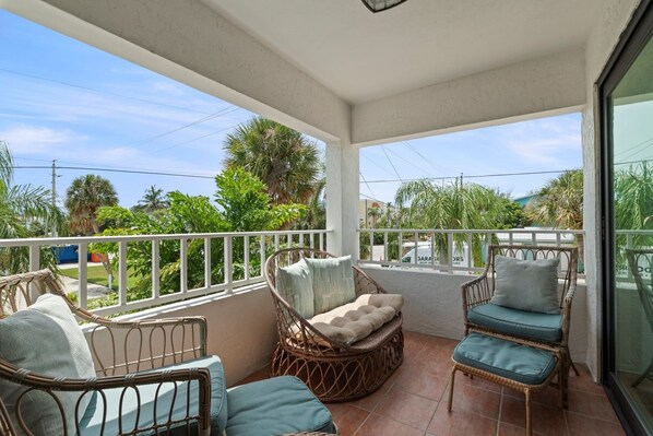 A balcony with wicker furniture, a white railing, and a view of palm trees and buildings.