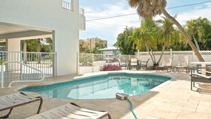 A swimming pool with lounge chairs and a table, surrounded by a white fence and palm trees. - Hidden Pearl 2 Bedroom Home by RedAwning (Holmes Beach)
