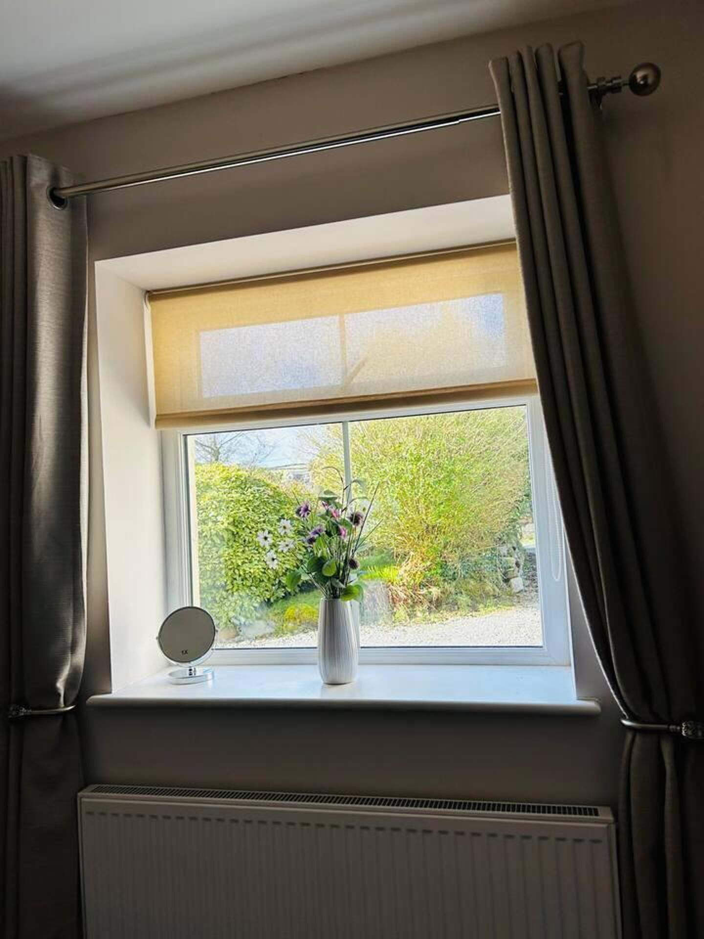 Una ventana con vistas a zonas verdes, un radiador debajo y un espejo redondo pequeño en el alféizar.