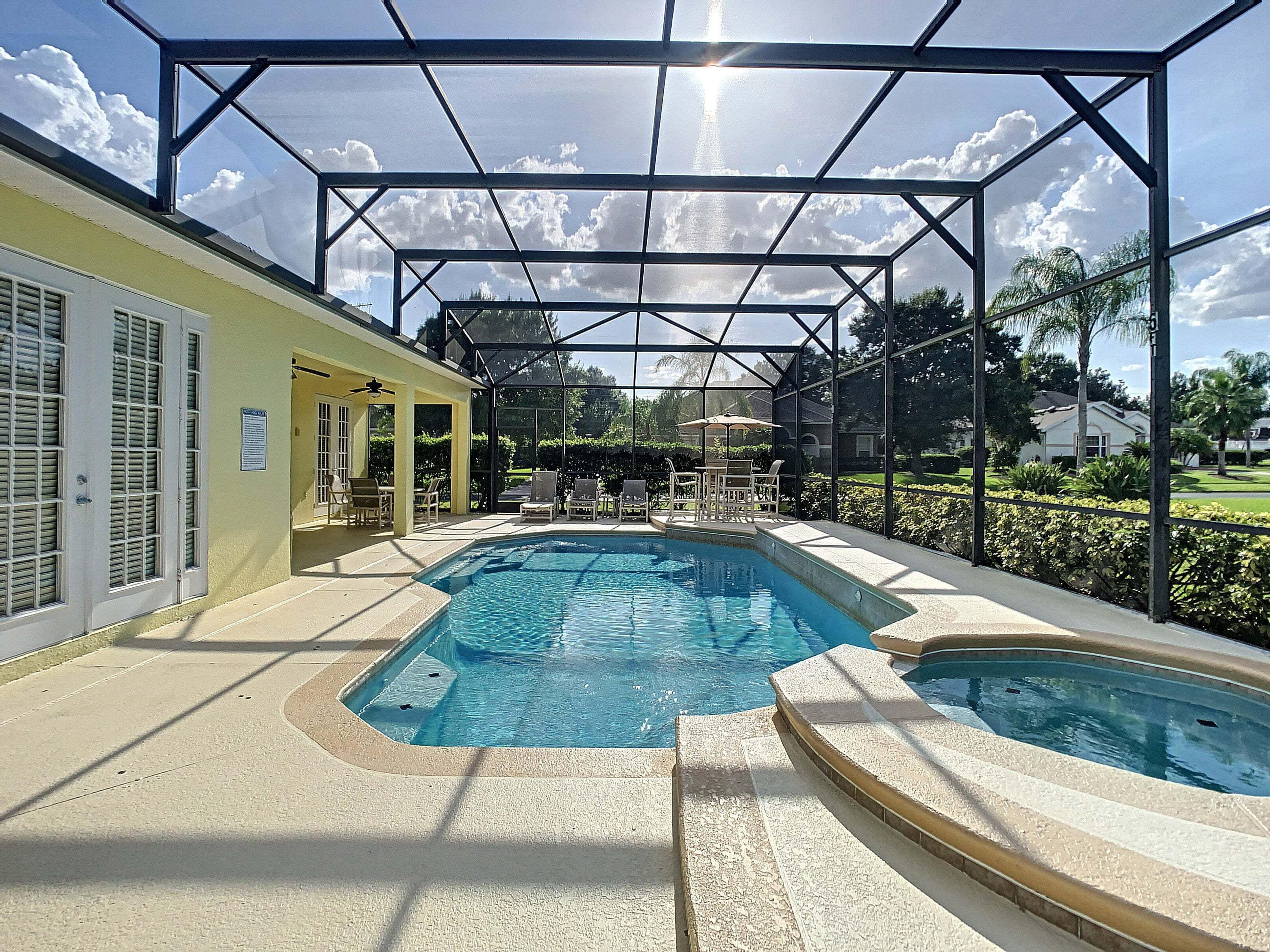 A swimming pool with a covered area and lounge chairs, surrounded by a fence and greenery.