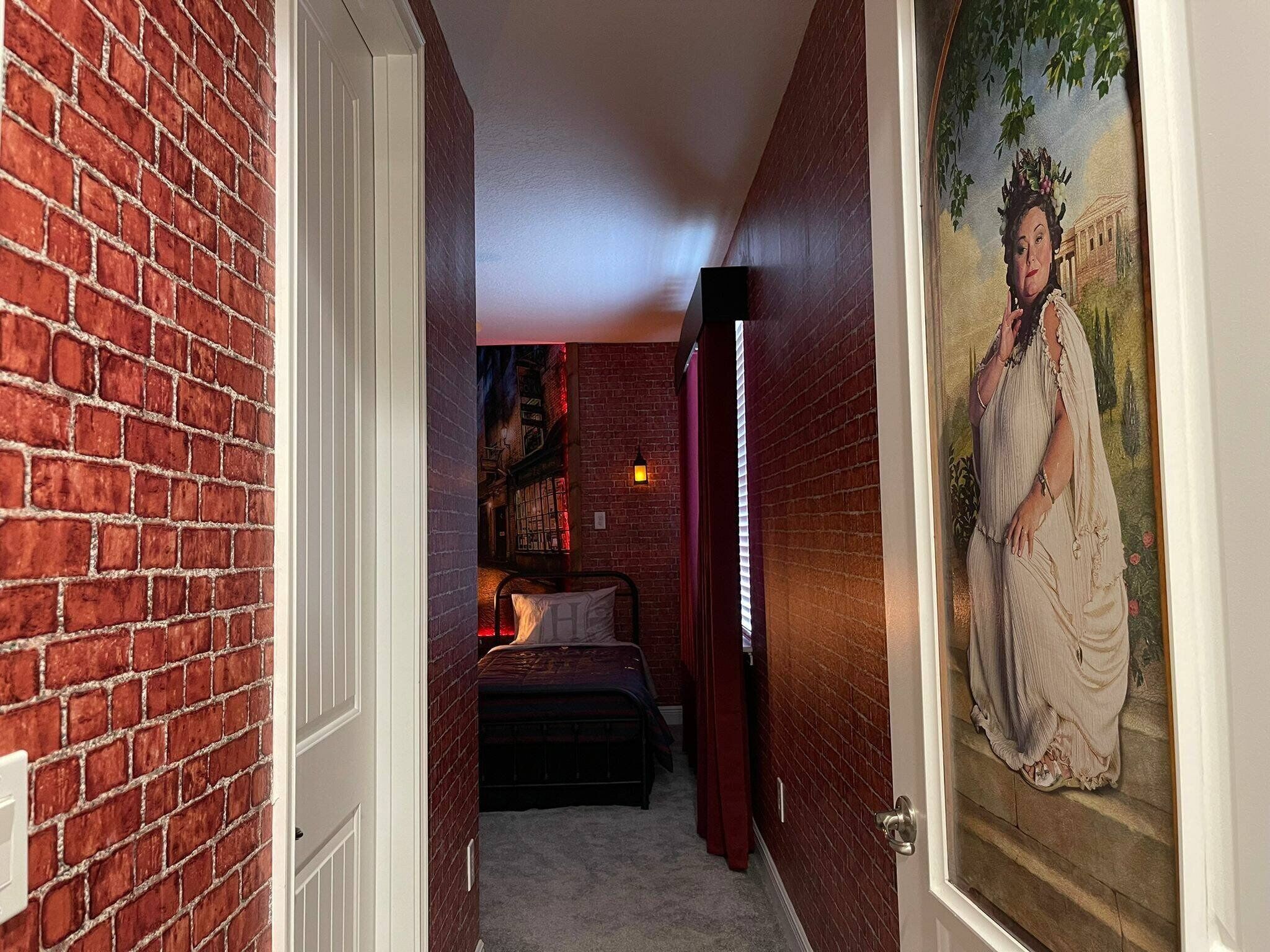 A narrow hallway with a brick wall on one side and a door on the other. There is a bed with a headboard and pillows in the room at the end of the hallway.