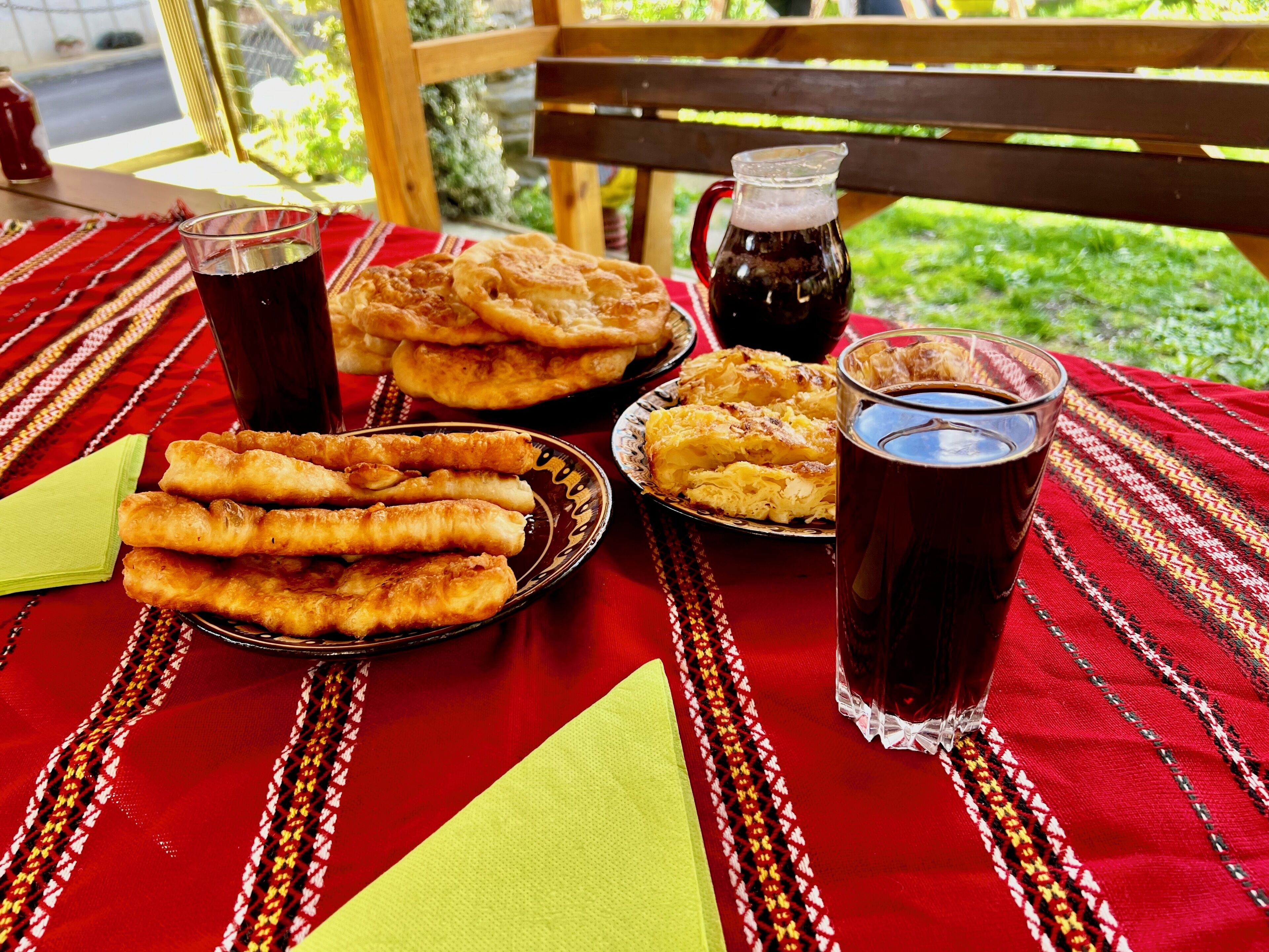 Une table garnie de plats et de boissons, notamment des pâtisseries frites et une carafe contenant un liquide sombre, probablement du café ou du thé, le tout posé sur une nappe rouge et jaune à motifs.
