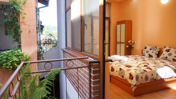 A bedroom with a bed, a window with blinds, a wooden door, and a balcony with a railing. - Pamir Guest House (Troyan)