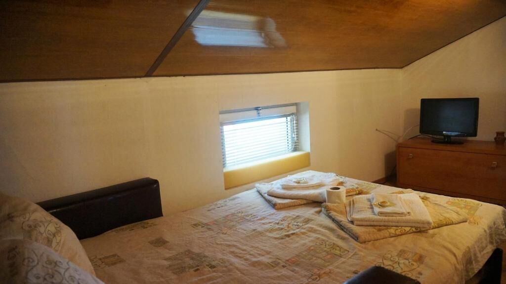 A small, cozy room with a single bed, a window with blinds, a television on a wooden dresser, and a sloped ceiling.