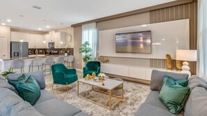Una sala moderna con un sofá gris, sillones turquesa y una mesa de centro. Se ve una cocina al fondo con un refrigerador y una zona de comedor.