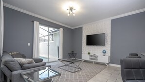 Un salon moderne avec un canapé gris, une table basse en verre, un téléviseur à écran plat fixé au mur et une porte-fenêtre coulissante donnant sur un balcon.