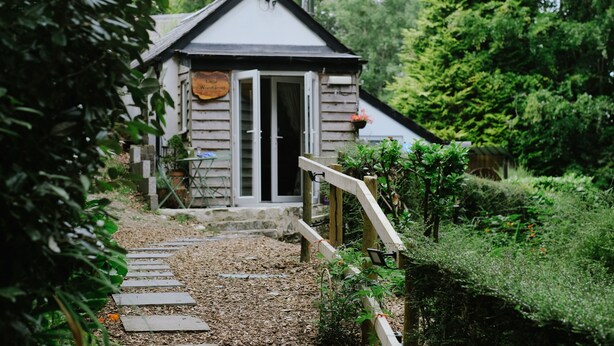 A rustic wooden building with a signboard, a wooden staircase leading to the entrance, and a garden path. - woodland cottage (barnstaple)