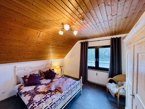 Ein Schlafzimmer mit Holzboden, einem Bett mit gemusterten Betttextilien, einer Nachttischlampe, einem Fenster mit Vorhängen und einem Sessel mit Felldecke.
