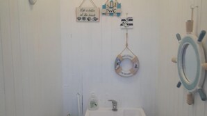 Un baño con lavamanos, inodoro y ducha. Hay elementos decorativos en la pared, incluyendo un bozal de salvamento y un letrero que dice 'Life is better at the beach'.