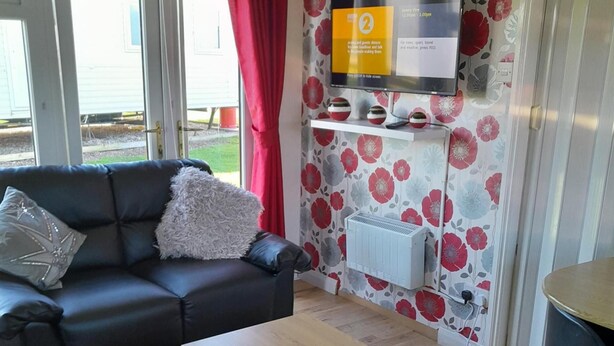 Una sala de estar con un sofá negro, un almohadón blanco y mullido, una mesa de centro de madera y un televisor en una pared con estampado floral.