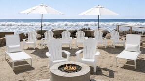 Un espace aménagé près de la plage, avec des chaises Adirondack blanches et des parasols, un foyer et une vue sur l’océan.