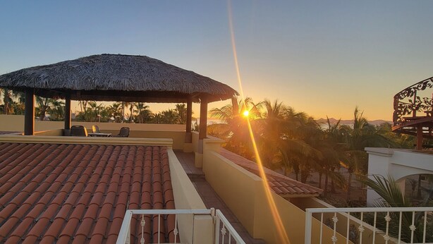 A rooftop terrace with a thatched roof structure, outdoor seating, and a view of palm trees and mountains during sunset.