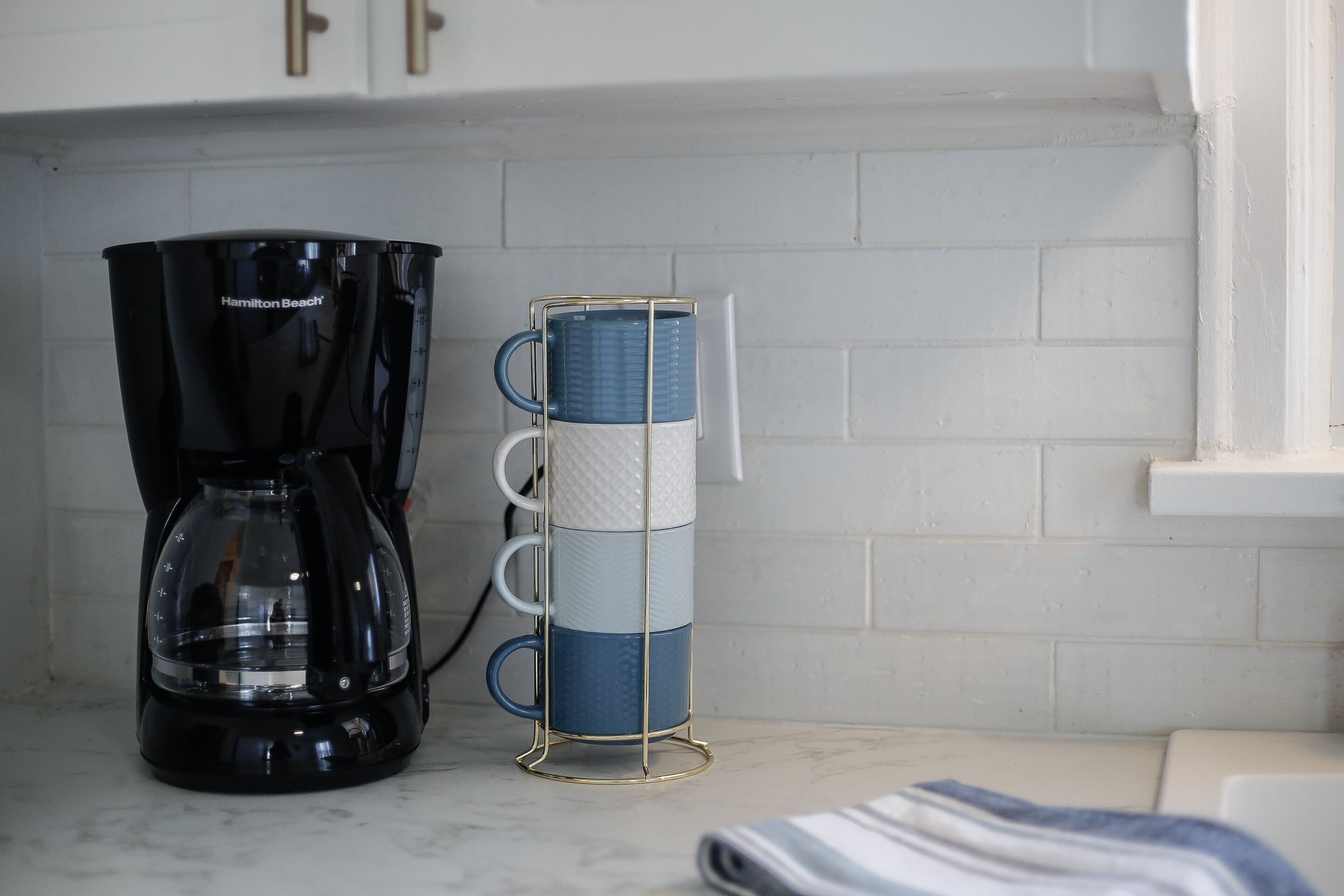 A kitchen with a black coffee maker and a stack of blue and white mugs on a white countertop.