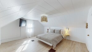 Une chambre avec un plafond en pente, un lit doté d’une tête de lit blanche, une table de chevet avec une lampe et une fenêtre ornée de rideaux légers.