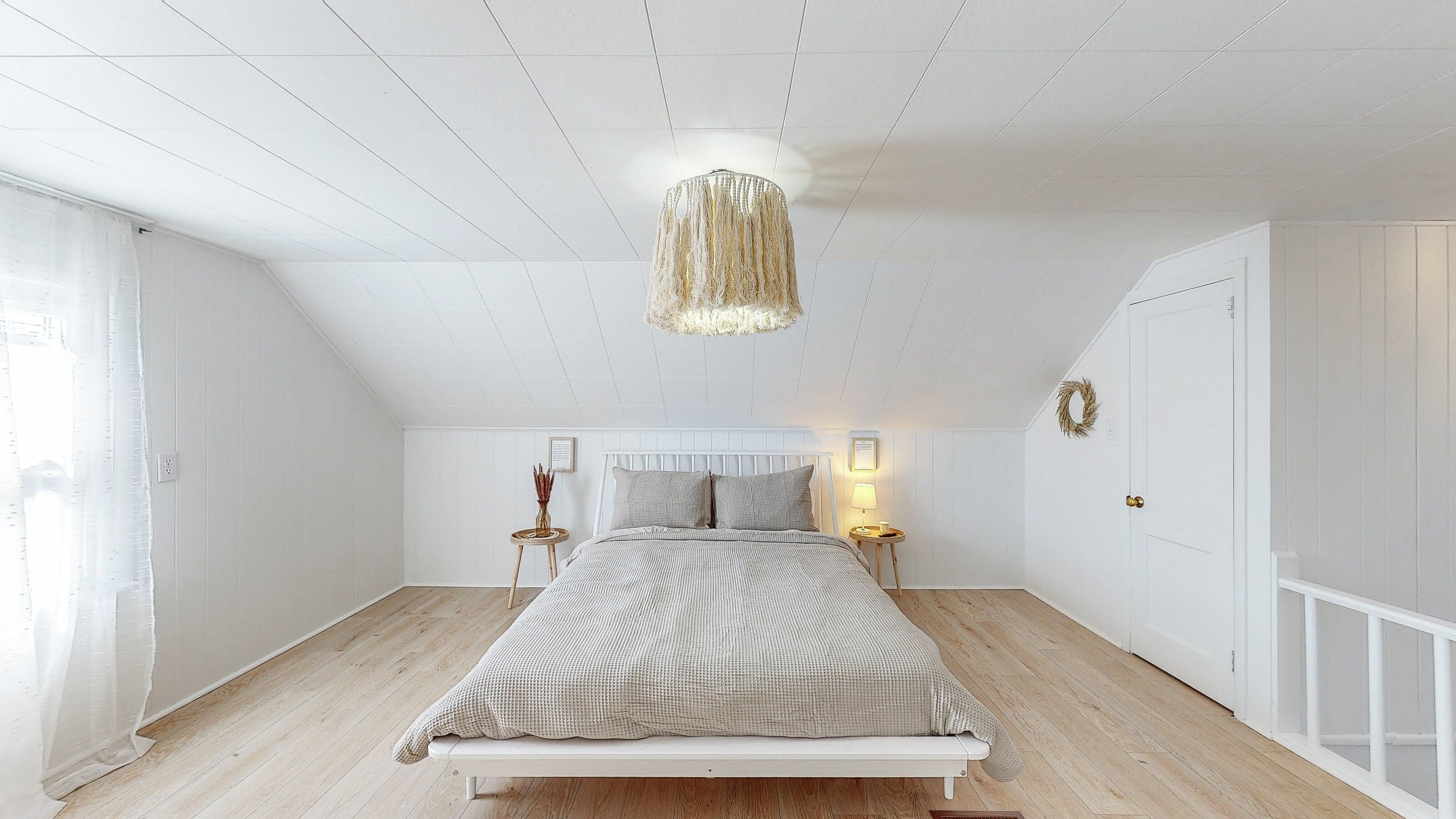 Un dormitorio con una cama blanca, un colgante de luz y una mesita de noche pequeña con una lámpara.