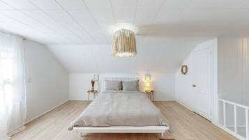 Un dormitorio con una cama blanca, un colgante de luz y una mesita de noche pequeña con una lámpara.
